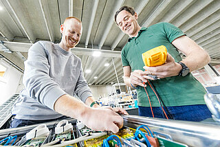 Two men, one with a voltmeter in his hand, stand in front of the assembled battery system and carry out the final inspection.